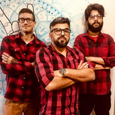 Marcos Sasso, Chevrand e Cadu com blusas xadrez vermelho e preto e braços cruzados no escritório da VX.